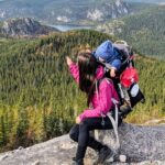Hiker wearing a pink jacket with a child in a backpack on a rocky overlook above a forested valley and distant mountains.