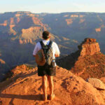 Hiker stands on a rocky ledge overlooking the Grand Canyon at sunset, wearing a backpack and shorts on a warm, arid overlook.