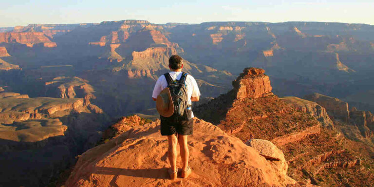 Hiker stands on a rocky ledge overlooking the Grand Canyon at sunset, wearing a backpack and shorts on a warm, arid overlook.