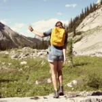 Hiker with an orange backpack standing on a rocky trail in a mountain meadow, mountains in the distance (back view).