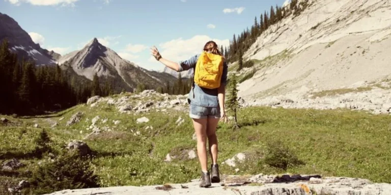 Hiker with an orange backpack standing on a rocky trail in a mountain meadow, mountains in the distance (back view).