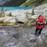 A hiker in a red backpack crosses a rocky stream with trekking poles, turquoise lake and mountains in the background.