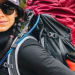 Person wearing a black long-sleeve top and sunglasses with a large hiking backpack outdoors, showing an orange water bottle in a side pocket.