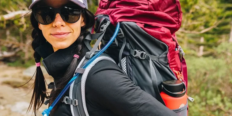 Person wearing a black long-sleeve top and sunglasses with a large hiking backpack outdoors, showing an orange water bottle in a side pocket.