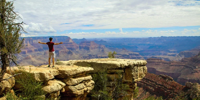 Person standing on a rocky ledge with arms outstretched, overlooking the Grand Canyon and vast canyon walls under a cloudy sky.