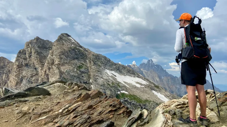 Hiker wearing an orange cap and backpack stands on a jagged rocky ridge with snow patches and mountain peaks in the distance under a cloudy sky.