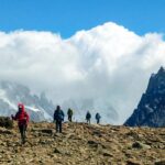 Group of hikers trekking a rocky mountain trail with snow-capped peaks and clouds in the background.