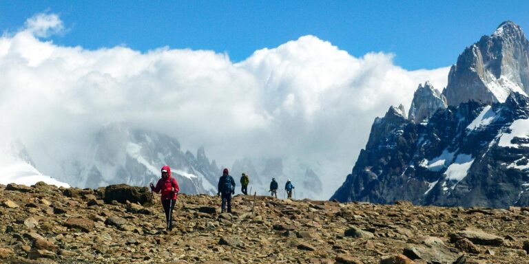 Group of hikers trekking a rocky mountain trail with snow-capped peaks and clouds in the background.