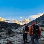 Two hikers with backpacks trek across a rocky, barren terrain toward snow‑capped mountains bathed in sunrise light.