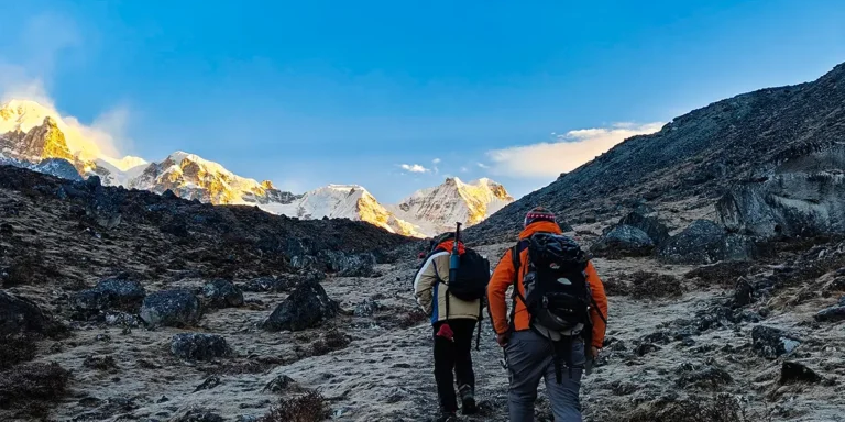 Two hikers with backpacks trek across a rocky, barren terrain toward snow‑capped mountains bathed in sunrise light.