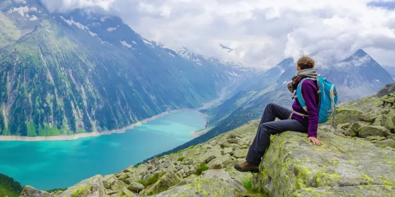 Hiker sitting on a mossy rock with a blue backpack, overlooking a turquoise alpine lake and towering green mountains.