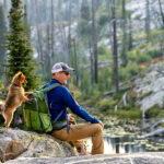 Hiker in a blue jacket and cap sits on a rock by a forest lake with a green backpack, a small dog standing on its hind legs beside him.