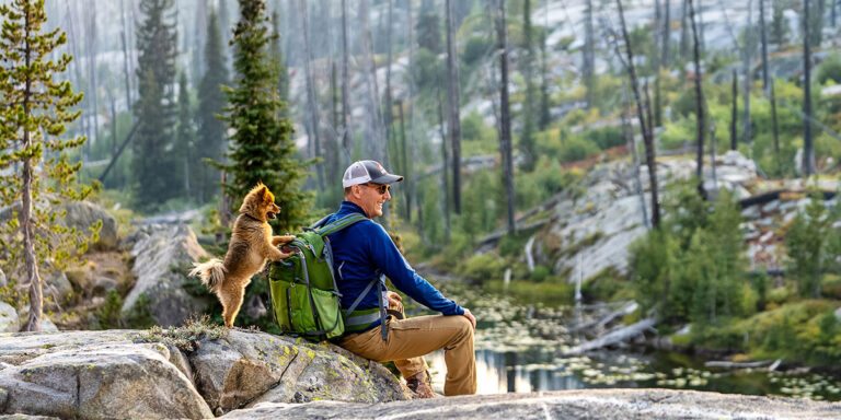 Hiker in a blue jacket and cap sits on a rock by a forest lake with a green backpack, a small dog standing on its hind legs beside him.