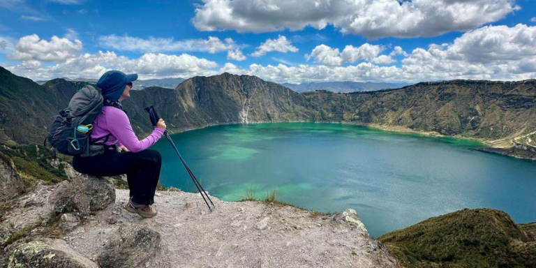 Hiker with a purple long-sleeve shirt sits on a rocky cliff overlooking a turquoise crater lake surrounded by rugged mountains.